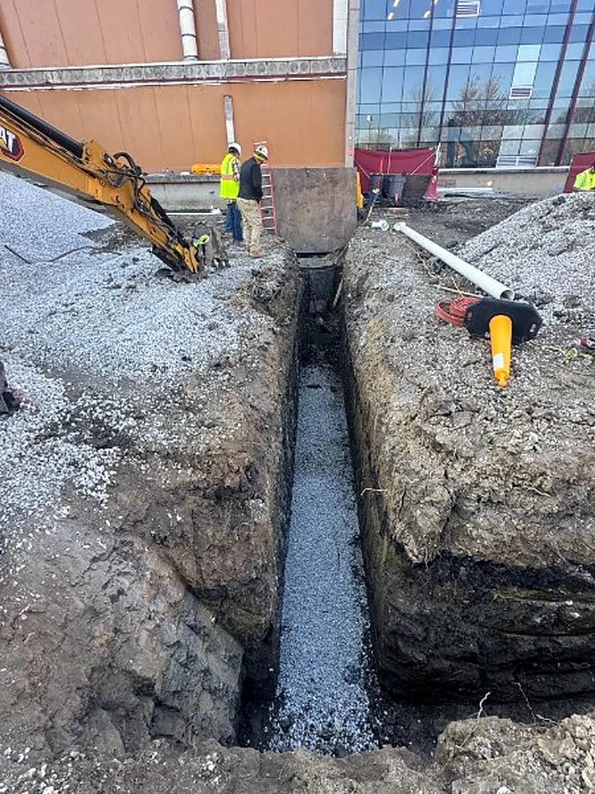 Excavator digging a deep utility trench beside a building with the crew on site